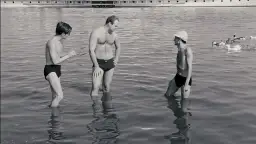 Trzech mężczyzn w kąpielówkach stoi w wodzie. Jeden z nich, Stanisław Tym w filmie Rejs, rozmawia z kolegami.