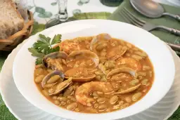 Plato de verdinas con almejas y gambas, servido en un cuenco blanco con perejil. Acompañado de pan en una cesta.