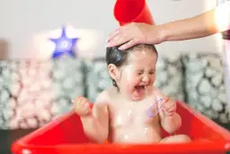 Małe dziecko panicznie boi się mycia głowy, krzyczy i płacze, gdy mama polewa mu włosy wodą z czerwonego kubeczka.