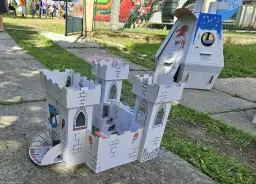 Zabawki z kartonu diy: biały zamek z wieżami i schodami, obok rakieta. Dzieci bawią się w tle.