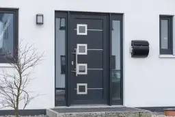 Nowoczesne drzwi zewnętrzne w ciemnoszarym kolorze z geometrycznymi przeszkleniami i metalowymi elementami. Obok skrzynka na listy.