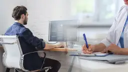 Mężczyzna pracuje przy komputerze, podczas gdy lekarz wypełnia dokumenty. Czy na L4 można pracować? To pytanie nurtuje wielu.