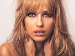 Kobieta z blond włosami i cieniowaniem grzywki, z dłońmi na szyi.