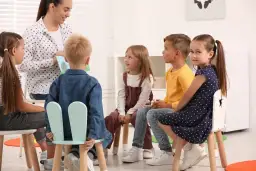 W przedszkolu nauczycielka czyta książkę grupie sześciorga dzieci. Wszyscy siedzą na krzesłach, słuchając uważnie.