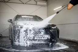 Czarny Lexus jest obficie spieniony podczas mycia na myjni bezdotykowej. Widać strumień wody z pianą, który opływa maskę i grill.
