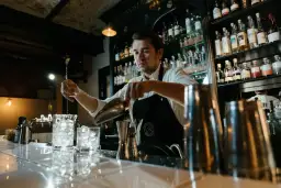 Barman przygotowuje drinka, zastanawiając się, ile zarabia dobry barman. Za nim półki z alkoholem.