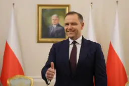 Mężczyzna w garniturze pokazuje kciuk w górę na tle portretu i flag Polski.