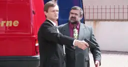 Gangster z filmu Kiler, w garniturze, celuje z pistoletu. Obok stoi mężczyzna w czarnym garniturze.