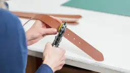 Ręce z dziurkaczem do skóry tworzą nową dziurkę w brązowym pasku.