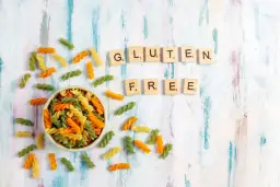 Kolorowy makaron w miseczce i rozsypane po stole, litery układające się w napis "GLUTEN FREE".