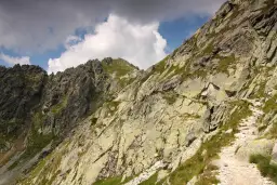 Kamienista ścieżka wije się po zboczu górskim, prowadząc w stronę skalistych szczytów. Widok na Szpiglasową Przełęcz, gdzie chmury przecinają błękit nieba.