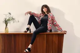Mujer con rizos sentada en mueble de madera, luciendo distintos tipos de estilos de vestir: chaqueta floral, camiseta con brillos y jeans negros.