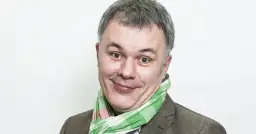 Uśmiechnięty mężczyzna w kratkowanym szaliku, znany jako Mazurek z RMF FM, z szarymi włosami i brązową marynarką.