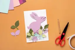 Uroczy różowy króliczek z papieru na kartce wielkanocnej DIY, otoczony kolorowymi kwiatami i liśćmi. Obok nożyczki, ołówek i taśma.