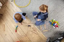 Dzieci budują tory kolejowe, angażując się w odgrywanie ról. Na dywanie leżą klocki, a obok drewniana kolejka.
