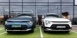 Dwa nowe samochody Kia Niro, jeden niebieski, drugi biały, stoją obok siebie na zielonym parkingu przed nowoczesnym budynkiem z dużymi oknami.