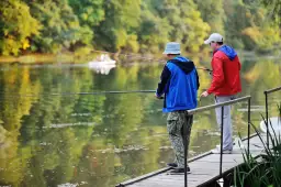 Dwóch mężczyzn łowi ryby z drewnianego pomostu na tle zielonego lasu i spokojnej wody.