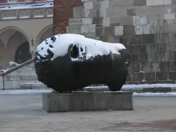 Duża, ciemna rzeźba głowy pokryta śniegiem, na tle kamiennej ściany i arkadowego budynku.