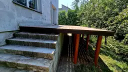 Nowy, wysoki taras na słupach, z drewnianą podłogą, sąsiaduje z betonowymi schodami i zielonym ogrodem.