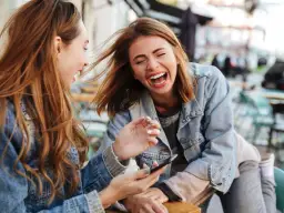 Dwie młode kobiety śmieją się na zewnątrz, jedna pokazuje coś na telefonie.