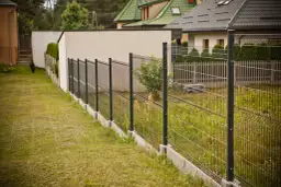 Nowoczesne ogrodzenie panelowe pod kątem wyznacza granicę posesji, za nim widać domy i zieleń.