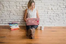Kobieta siedzi na podłodze, pracując na różowym laptopie. Obok stos książek i kubek. Uczy się pilnie, jak pisze się zręcznie.