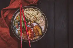 A miseczce z aromatycznym ramen wegetariańskim, pełnej makaronu, grillowanego bakłażana i pomidorów, spoczywają czerwone pałeczki.