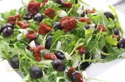 Sałatka z rukolą, czarnymi oliwkami, suszonymi pomidorami i kawałkami mozzarelli, polana dressingiem.