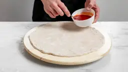 Ręce dodają sos pomidorowy do surowego ciasta na pizzę na drewnianej desce.
