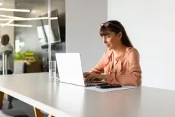 Kobieta pracuje przy laptopie, przygotowując rachunek do umowy o dzieło. Na biurku stoi szklanka wody i telefon.