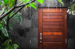 Deszcz spływa po pięknych, drewnianych drzwiach zewnętrznych dębowych, obok których rośnie bujna, zielona roślinność.
