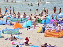 Tłum ludzi na plaży, opalający się i kąpiący w morzu, kolorowe parawany i namioty plażowe.