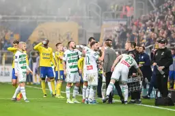 Piłkarze w białych i żółtych strojach piją wodę na boisku, w tle kibice.