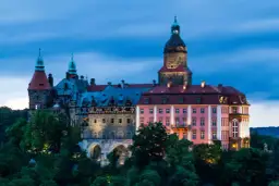 Zamek Książ o zmierzchu, z różową fasadą i kamiennymi wieżami, otoczony zielenią.