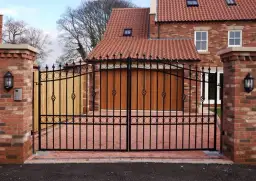 Kuta brama wjazdowa z czarnego metalu, ozdobiona ornamentami, przed domem z czerwonej cegły.