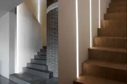 Nowoczesne schody z drewnianymi stopniami i szarymi stopniami, oświetlone pionowymi ledami w ścianie przy schodach.