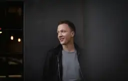 Joris, lächelnd, lehnt an einer dunklen Wand.