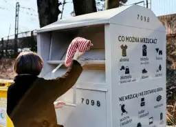 Kobieta wrzuca ubrania do szarego kontenera na odzież. Na kontenerze widnieje napis "CO MOŻNA WRZUCAĆ?".