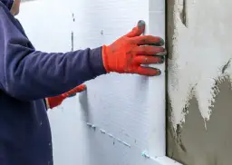 Ręce w pomarańczowych rękawicach przyklejają biały styropian do ściany piwnicy.