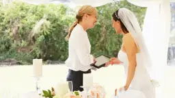 Panna młoda z welonem i wedding plannerka dyskutują przy stole. Czy warto wynająć wedding plannera, czy poradzić sobie samemu?