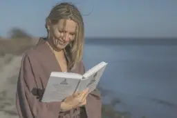 Kobieta w brązowym kimonie czyta książkę na plaży, z morzem w tle.