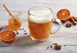 Kubek piwa z pianką, obok plasterki pomarańczy, miód, cynamon i goździki.
