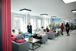 Grupa seniorów siedzi na kanapach w jasnym, nowoczesnym wnętrzu domu opieki.