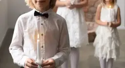 Dziecko w białej koszuli z czarną muszką trzyma zapaloną świecę. W tle inne dzieci w białych strojach.