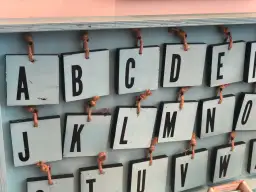 Drewniane tabliczki z literami alfabetu angielskiego pisanego, wiszące na sznurkach. Widoczne litery A, B, C, D, E, J, K, L, M, N, O, U, V, W.
