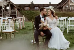 Para młoda siedzi na krzesłach na tle drewnianych budynków i białych krzeseł.