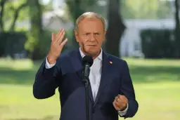 Donald Tusk w garniturze, przemawiający do mikrofonu, z serduszkiem na klapie.