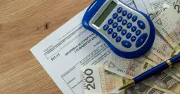 Formularz PIT-11, kalkulator, długopis i polskie banknoty na drewnianym tle.