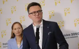 Szymon Hołownia w okularach i garniturze przemawia przy mikrofonie, za nim kobieta i logo Polska 2050.