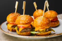 Mini burgery z serem, pomidorem i sałatą na talerzu, ozdobione patyczkami.
